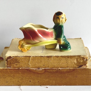May include: A vintage ceramic planter in the shape of a boy pushing a flower cart. The cart is a tulip with pink and yellow petals. The boy is wearing a green outfit and has brown hair. The planter is sitting on top of old books.