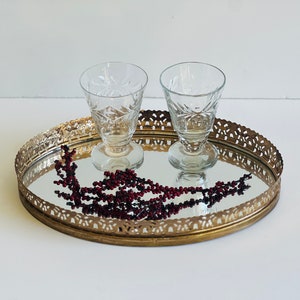 K&ouml;nnte beinhalten: Ein ovales, vergoldetes Spiegeltablett mit einem dekorativen Filigranrand. Zwei klare Schnapsgl&auml;ser mit ge&auml;tzten Designs befinden sich auf dem Tablett. Ein Strang roter Beeren liegt auf dem Tablett.