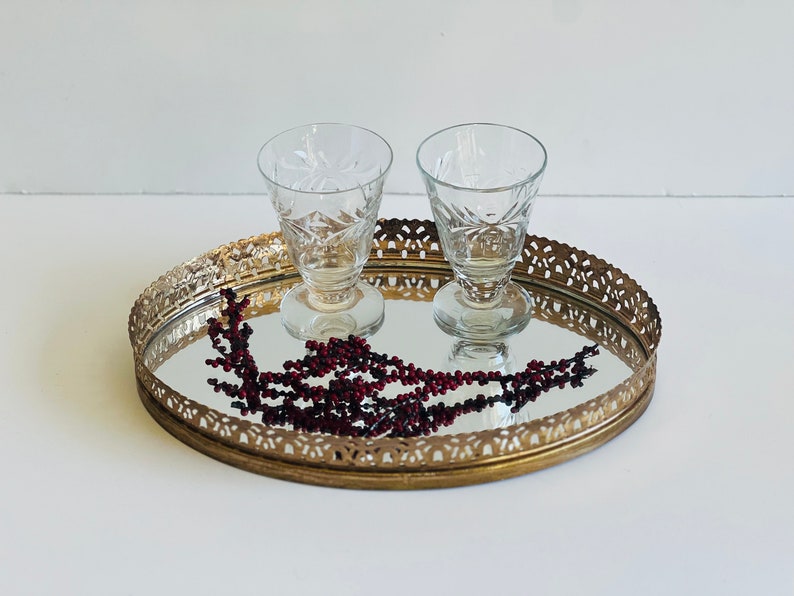 K&ouml;nnte beinhalten: Ein ovales, vergoldetes Spiegeltablett mit einem dekorativen Filigranrand. Zwei klare Schnapsgl&auml;ser mit ge&auml;tzten Designs befinden sich auf dem Tablett. Ein Strang roter Beeren liegt auf dem Tablett.