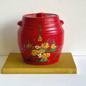 Puede incluir: Un tarro de cerámica rojo con tapa y dos asas pequeñas. El tarro está decorado con diseños florales amarillos y verdes. Se encuentra sobre un libro amarillo. El tarro mide unos 15 cm de alto.