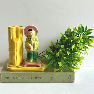 May include: A vintage yellow ceramic vase with a bamboo-like design, next to a figurine of a person in a hat and green clothing. The figurine is holding an object. The items are on a green book with the text "THE ACHIEVING SOCIETY".