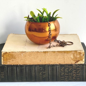 May include: A small, round copper-colored hanging planter filled with green artificial plants. The planter hangs from a chain and sits atop an antique book. The book has aged, yellowed pages and is stacked on top of another dark book.