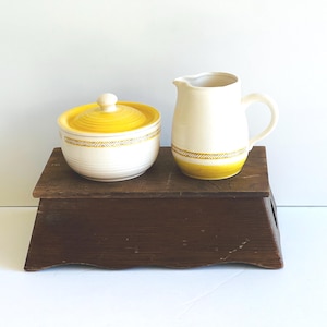 May include: A vintage cream and yellow ceramic sugar bowl with lid and a matching pitcher. The sugar bowl and pitcher have a yellow band with a geometric design. The items are displayed on a dark brown wooden stand.