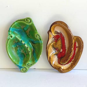 May include: Two ceramic ashtrays. One is leaf-shaped with green and blue glaze, accented with gold. The other is a shell-like shape with brown, red, and beige glaze. Both have a glossy finish and are on a white background.