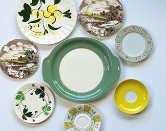 May include: An assortment of vintage ceramic plates and saucers in various sizes and designs. Includes a large green serving dish, a yellow saucer, and plates with floral and landscape patterns. The plates are arranged on a white surface.