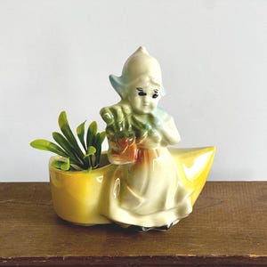 May include: A vintage ceramic planter in the shape of a Dutch girl holding a basket. The planter is painted in shades of yellow, cream, and blue, with a small compartment for plants. The planter is filled with green artificial plants.
