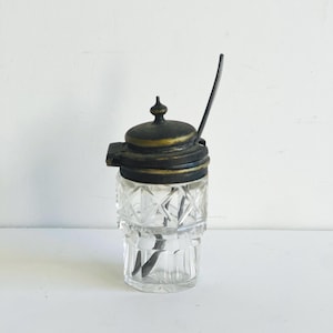 May include: A clear glass jar with a cut glass design and a black and gold metal lid. A small metal spoon is inside the jar.