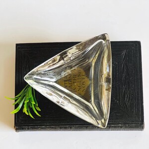 May include: A clear, triangular ashtray with rounded corners. The ashtray is made of glass and sits on a dark, textured surface. A sprig of green leaves is placed near the ashtray.