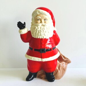 May include: A ceramic Santa Claus figurine in a classic red suit with a black belt and boots. The figurine has a white beard, a red hat, and a black glove. A brown sack is at his side, ready for Christmas.