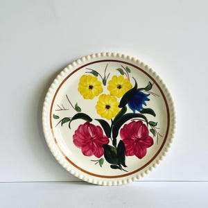 May include: Cream-colored ceramic plate with a scalloped edge and a brown rim. The plate features a hand-painted floral design with yellow, red, and blue flowers, along with green leaves. A decorative piece for serving or display.