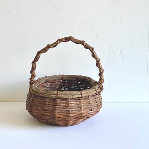 May include: A round woven basket crafted from brown willow branches, featuring a thick, textured rim and a single arched handle wrapped with a thinner, darker vine. The basket has a deep, open interior and a sturdy, circular base structure.