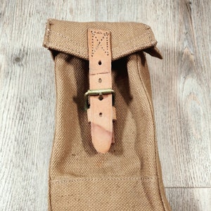 Peut inclure: Une pochette en toile beige avec une sangle en cuir et une boucle en laiton. La pochette est de forme rectangulaire avec un rabat supérieur. La sangle en cuir est fixée par des coutures et des rivets. Fond en bois.