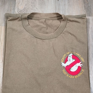 May include: A brown t-shirt with an embroidered patch featuring a ghost in a red circle with the text "ECMFPTF HI-KABUL 2007 THAT'S HOW WE ROLL III"