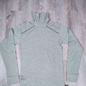 Puede incluir: Una camiseta de cuello alto de manga larga de color verde claro con un patrón texturizado. La camiseta tiene una pequeña etiqueta negra y dorada en la parte inferior derecha.