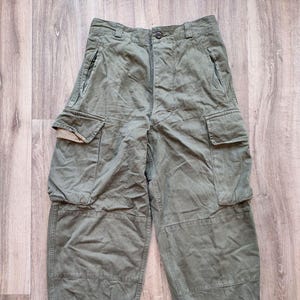 May include: Olive green cargo trousers with multiple pockets and a button closure. The trousers have a relaxed fit with gathered ankles. The fabric appears to be a durable, possibly military-style material.
