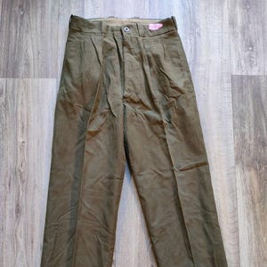 Puede incluir: Pantalones verde oliva con cierre de botón y pliegues. Los pantalones de cintura alta tienen un diseño de pierna recta. La tela parece ser de peso medio. Una etiqueta rosa está adherida a la cintura.