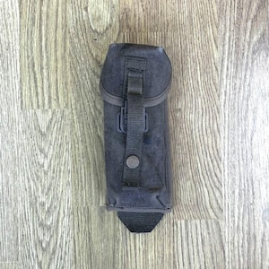 以下が含まれることがあります： 木製の表面に置かれた、フラップ留めとバックル付きストラップが付いた黒いキャンバスポーチ