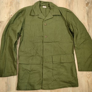 Puede incluir: Una chaqueta de trabajo verde con cuatro botones y dos bolsillos grandes. La chaqueta tiene cuello y mangas largas.