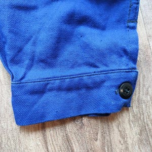 May include: Close-up of a bright blue fabric, possibly denim or a similar material. The image focuses on a buttoned cuff, showcasing the texture and colour of the garment. A black button is visible.