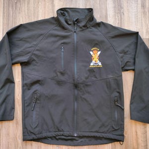 May include: A black softshell jacket with a full-length zip, a stand-up collar, and multiple zipped pockets. The jacket features an emblem on the chest with text that reads "Apotille Mountain Platoon".