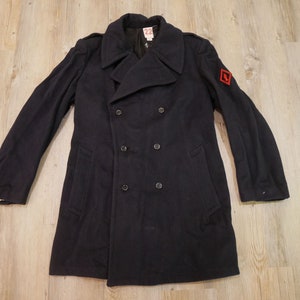 May include: A dark blue double-breasted peacoat with a red patch on the right sleeve. The coat has a high collar and two pockets on the front.