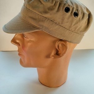 May include: A khaki-colored military-style cap with a short brim and two black eyelets on the side. The cap is made of a textured fabric and has a structured design. The cap is displayed on a mannequin head.