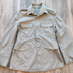 Può includere: Una giacca di colore marrone chiaro in stile militare con chiusura a bottoni e due tasche a patta. La giacca ha un colletto e maniche lunghe.