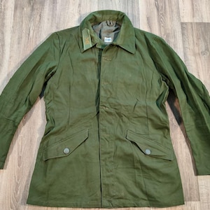 Puede incluir: Una chaqueta verde de estilo militar con cierre de botones y dos bolsillos de solapa. La chaqueta tiene cuello y mangas largas.