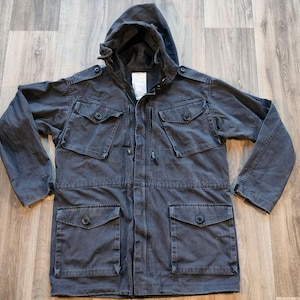 May include: A dark grey hooded jacket with multiple pockets and a zip closure. The jacket has a drawstring hood and a flap over the zip.