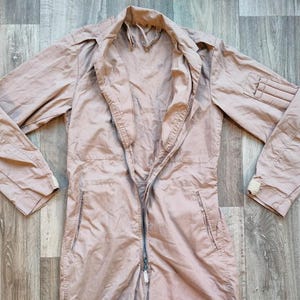 Peut inclure: Combinaison de couleur beige avec une fermeture éclair intégrale, des manches longues et plusieurs poches. La combinaison a un col et une coupe ajustée. Le tissu semble léger et adapté aux températures chaudes. Le vêtement est posé à plat sur une surface en bois.