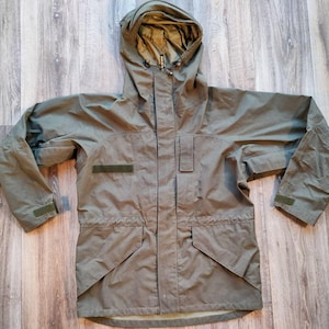 以下が含まれることがあります： オリーブグリーンのフード付きジャケット。フルレングスのジッパー、2つのフロントポケット、調節可能な袖口が付いています。左胸にパッチがあり、フードにはドローコードが付いています。耐久性のある防水素材で作られています。