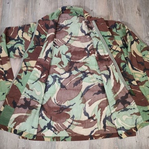 May include: A camouflage jacket with a green, brown and black pattern. The jacket is unzipped and laid flat, showing the inside lining.