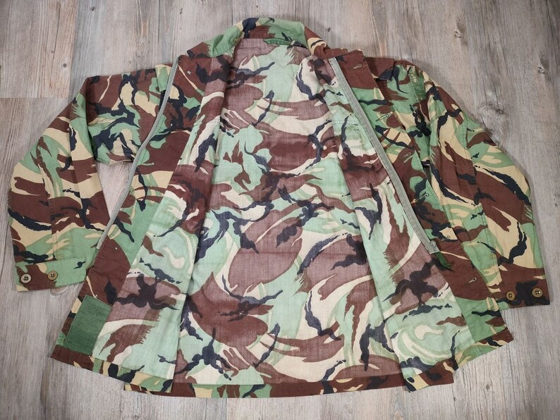 May include: A camouflage jacket with a green, brown and black pattern. The jacket is unzipped and laid flat, showing the inside lining.
