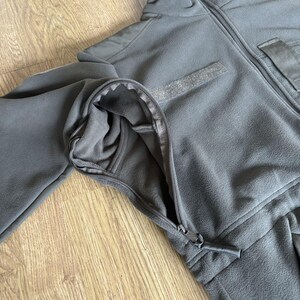 May include: A dark gray fleece jacket with a zipper closure and a pocket on the right side. The jacket has a high collar and a zippered pocket on the left sleeve.