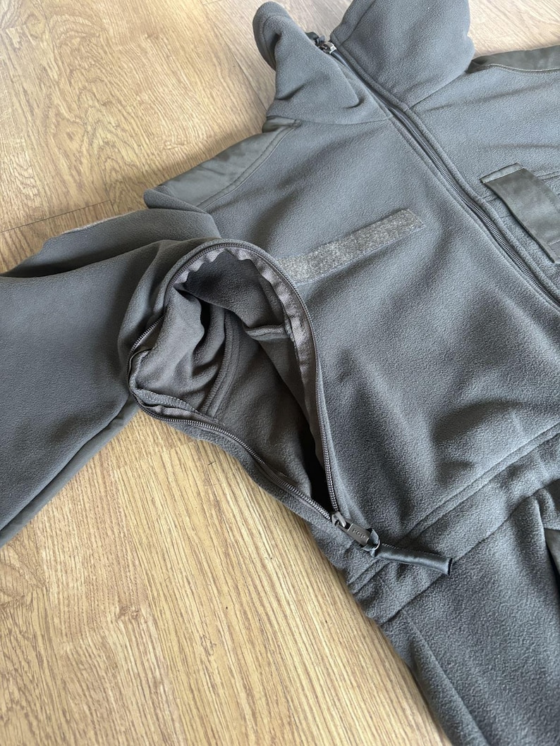 May include: A dark gray fleece jacket with a zipper closure and a pocket on the right side. The jacket has a high collar and a zippered pocket on the left sleeve.