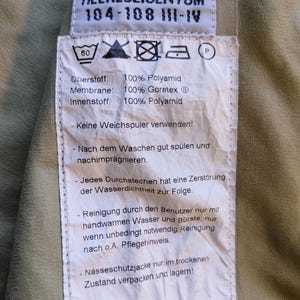 May include: A close-up of a tag with the text "HEERESEIGENTUM" and washing instructions. The tag is white with black text and is attached to a khaki-colored fabric. The tag also includes the text "100% Polyamid" and "100% Goretex".