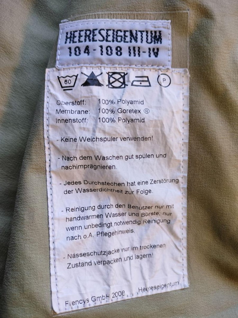 May include: A close-up of a tag with the text "HEERESEIGENTUM" and washing instructions. The tag is white with black text and is attached to a khaki-colored fabric. The tag also includes the text "100% Polyamid" and "100% Goretex".