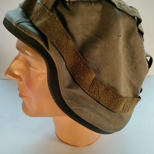 Puede incluir: Gorra de estilo militar verde oliva con visera negra. Cuenta con correas marrones ajustables y un aspecto desgastado y texturizado. La gorra se exhibe en una cabeza de maniquí.