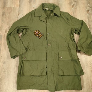 Peut inclure: Veste de style militaire vert olive avec un col, une fermeture boutonnée et deux grandes poches avant. Un écusson avec un blason est cousu sur la poitrine gauche. La veste est posée à plat sur une surface en bois.