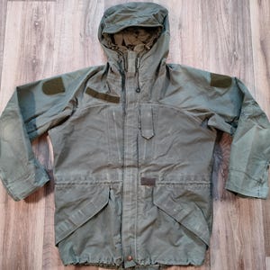 May include: A green hooded jacket with a zip closure and multiple pockets. The jacket has a drawstring hood and a flap over the zip.