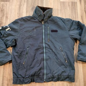 Puede incluir: Una chaqueta azul oscuro con cierre de cremallera y forro polar. La chaqueta tiene dos bolsillos en la parte delantera y dos bolsillos en las mangas. La chaqueta tiene un logotipo blanco en la manga izquierda.