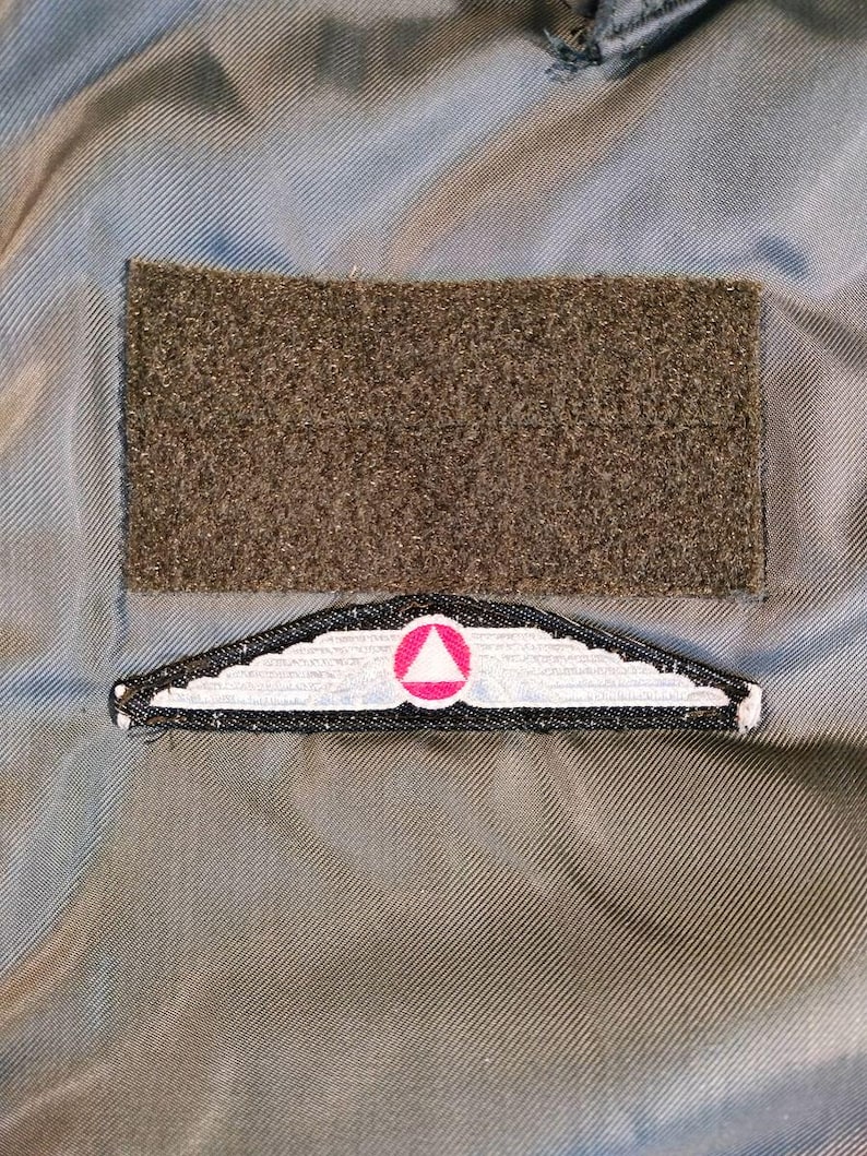 以下が含まれることがあります： 2 つのパッチが付いた軍服のクローズアップ。上部のパッチは、長方形のオリーブグリーンの面ファスナーです。その下には、ピンクの三角形と翼のデザインが施された白と黒のパッチがあります。