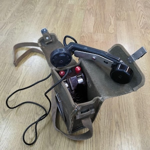 May include: A vintage green canvas bag with a black and silver phone handset inside. The phone has a black cord and two red buttons. The bag has a shoulder strap.