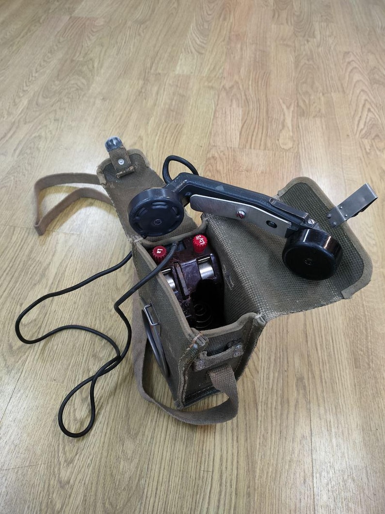 May include: A vintage green canvas bag with a black and silver phone handset inside. The phone has a black cord and two red buttons. The bag has a shoulder strap.