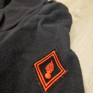 May include: A black wool jacket with a red embroidered patch featuring a grenade and a wing. The patch is on the left sleeve of the jacket.