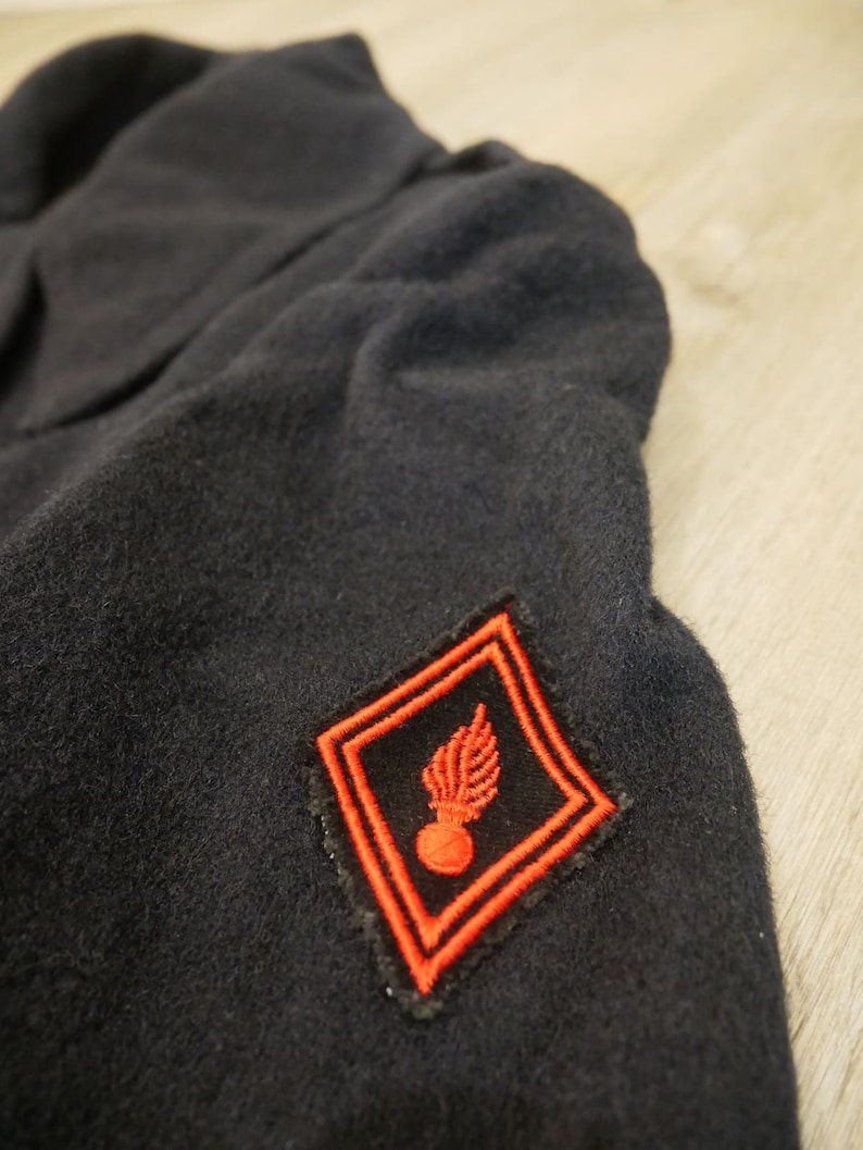 May include: A black wool jacket with a red embroidered patch featuring a grenade and a wing. The patch is on the left sleeve of the jacket.