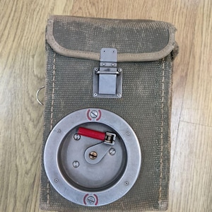 May include: A grey canvas bag with a metal clasp and a round metal spool with a red handle. The spool is attached to the bag.