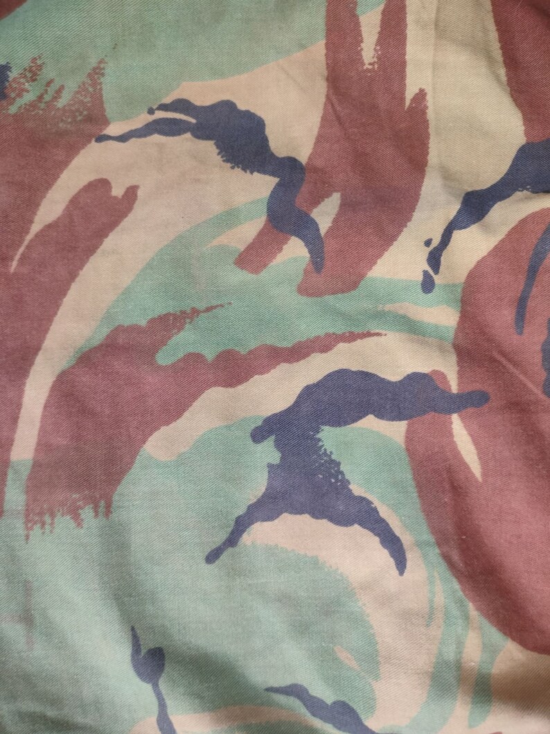 May include: Close-up of a camouflage fabric with a pattern of brown, green, and blue. The fabric has a textured surface and is made of a woven material.