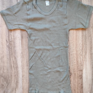 Peut inclure: Un t-shirt vert sauge en tricot côtelé à manches courtes. Le t-shirt a un col rond et une étiquette près de l'encolure. Le tissu est à côtes verticales. Le t-shirt est posé à plat sur une surface en bois.