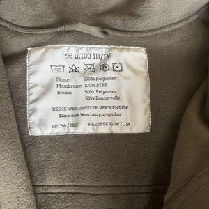May include: A white label with black text and symbols. The label reads "96 u.100 III/IV", "40&deg;", "Fleece: 100% Polyester", "Membrane: 100% PTFE", "Besatz: 50% Polyester 50% Baumwolle", "KEINE WEICHSP&Uuml;LER VERWENDEN!", "Nach dem Waschen gut sp&uuml;len", "FECSA/2017", "HEERESEIGENTUM".